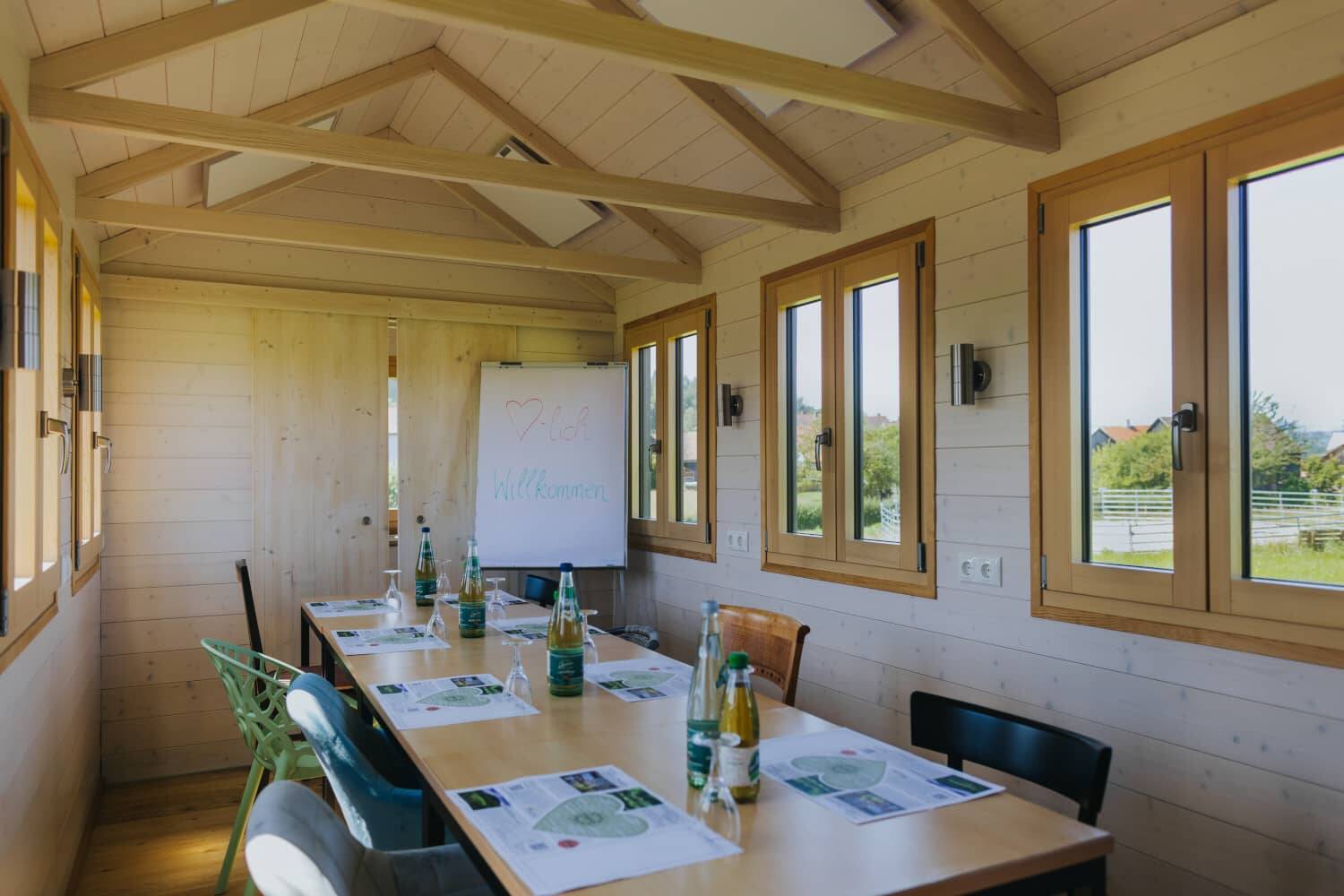 The interior of the Tiny House as a conference room, with light-coloured wooden walls and wooden table with conference documents and drinks. The window offers a view of the natural surroundings of the Hollerhöfe.