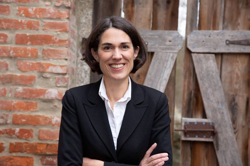 Astrid Köppel steht vor einer Holztür und lacht in die Kamera – Referentin beim Business-Frühstück in den Hollerhöfen mit Fokus auf persönliche Wirkung.