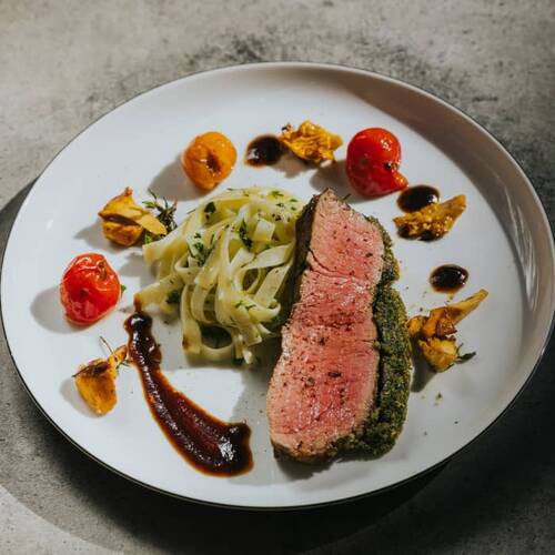 Fein angerichtetes Gericht mit rosa gebratenem Fleisch in Kräuterkruste, Tagliatelle, Cherrytomaten und dekorativen Saucenpunkten auf einem weißen Teller.