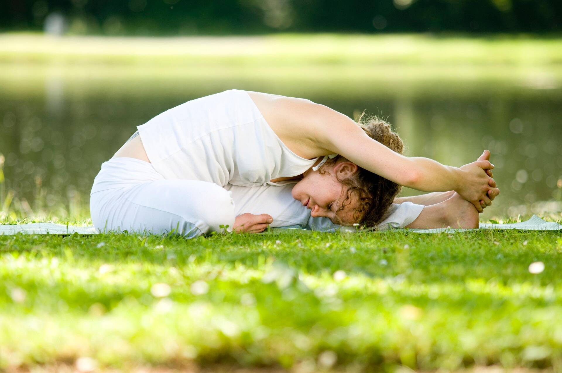 Woman in white yoga clothing sits in the greenery and practises a forward bend in nature; calm, natural atmosphere in the summer light.