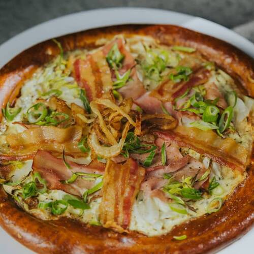 Herzhafter Flammkuchen mit Speck, Zwiebeln und Frühlingslauch auf einem weißen Teller – ein Beispielgericht aus den Hollerhöfen als Eventlocation mit Buffet.