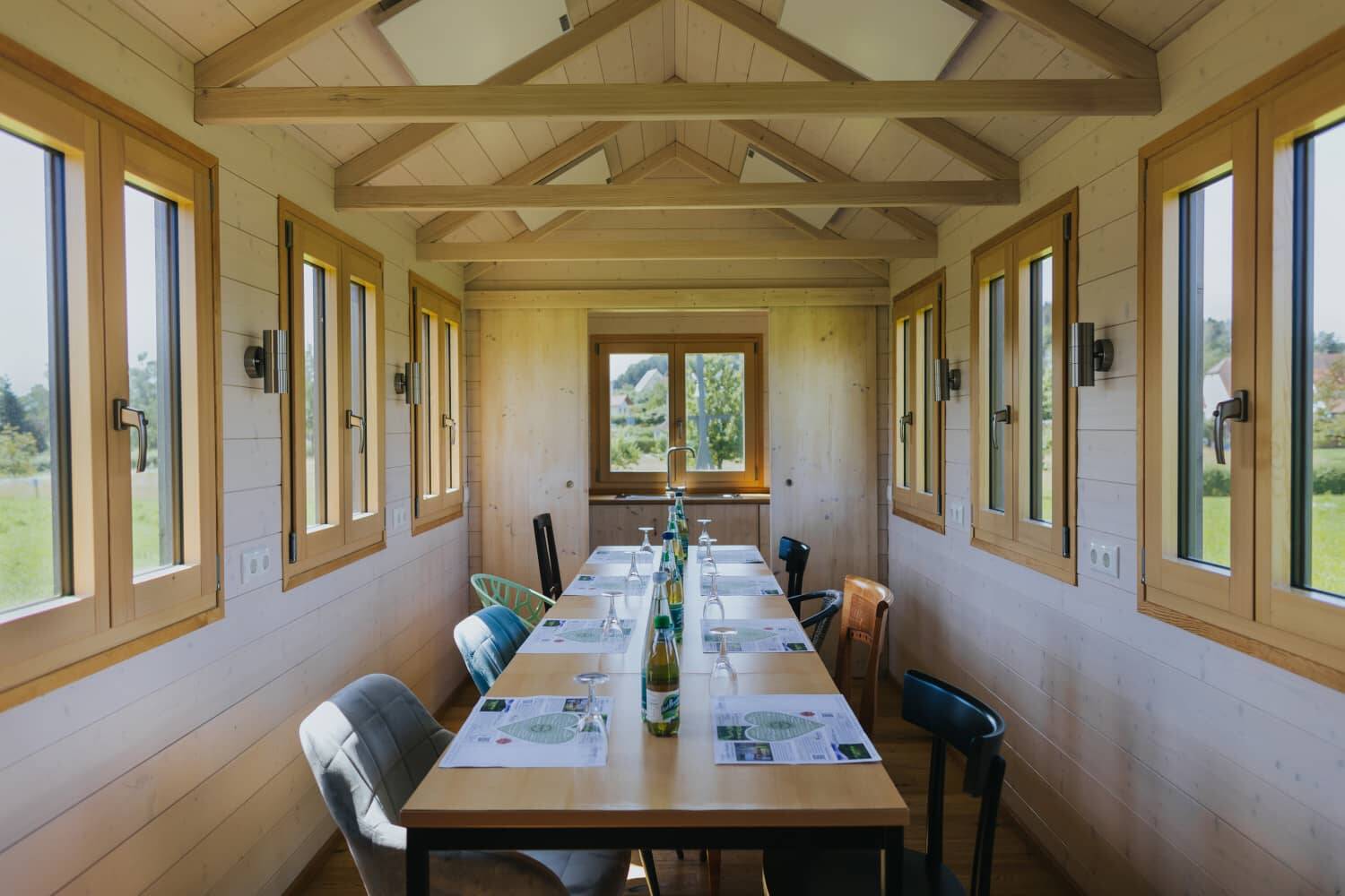 A symmetrical picture of the interior of the Tiny House, showing the conference table in the centre and three windows to the left and right with a view of the greenery.