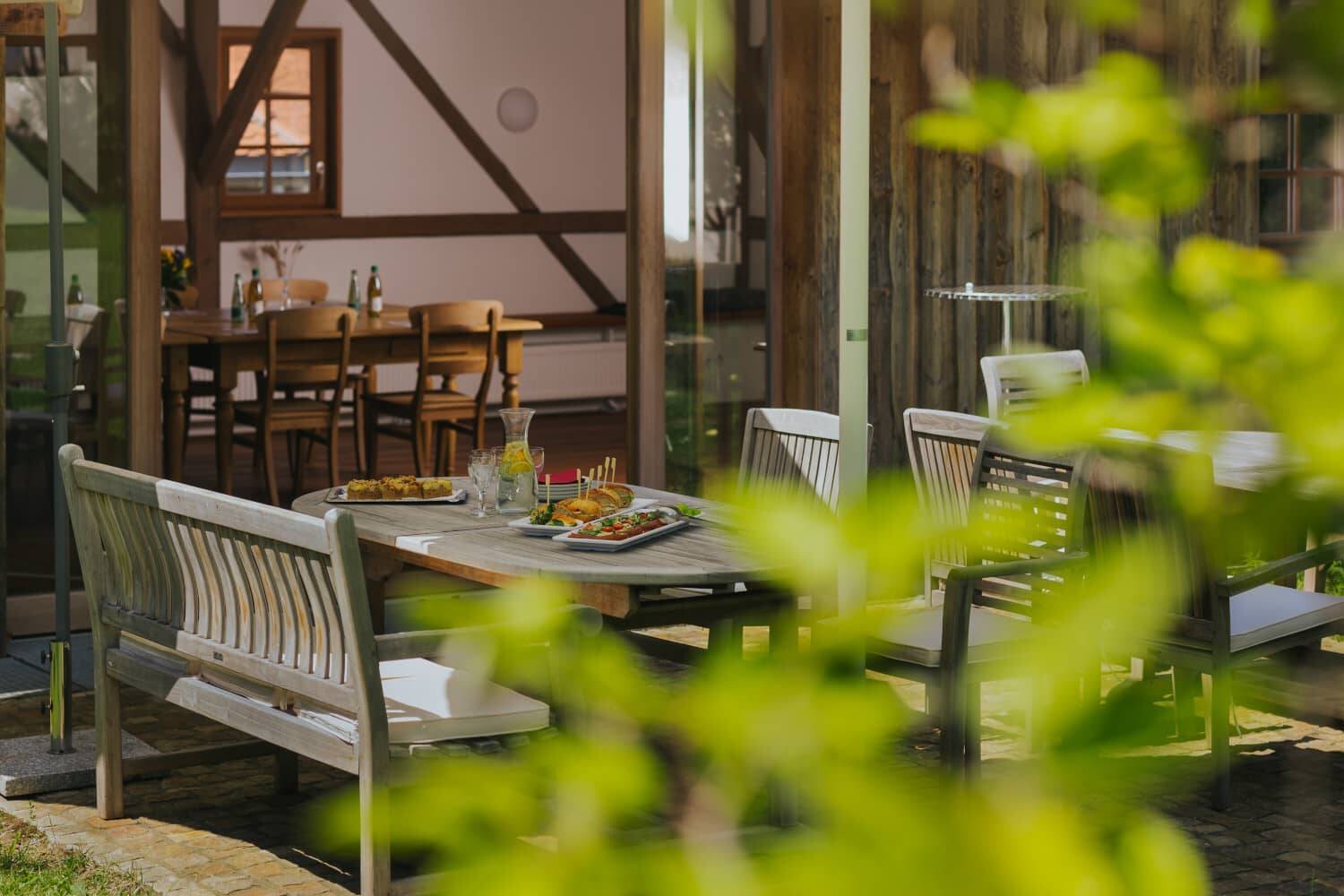 Atmospheric picture with blurred leaves in the foreground, behind it a set wooden table outside with delicious conference catering, snacks and fresh juice.