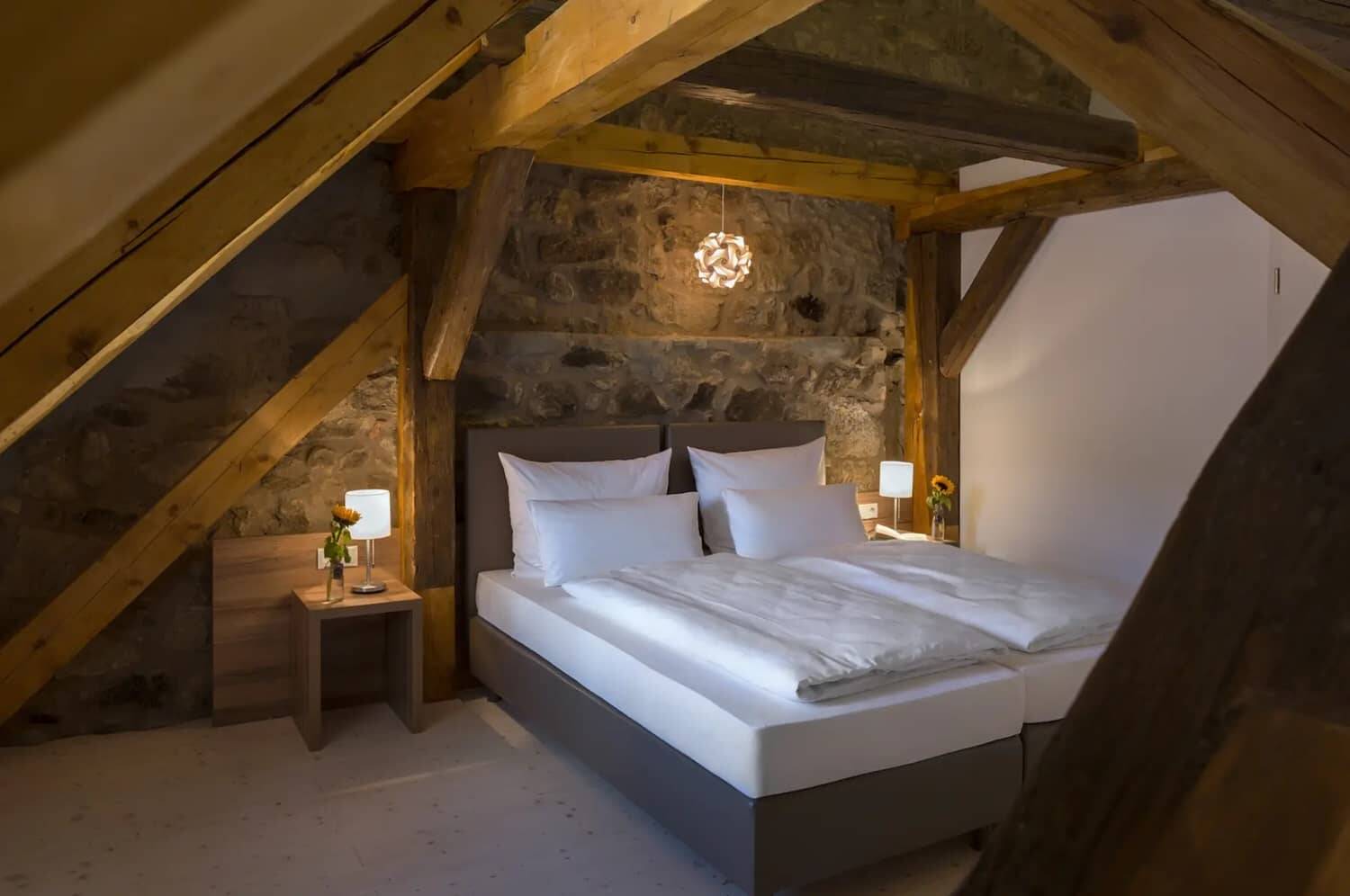 Das Bett im Schlafzimmer des Deluxe Apartments, rustikal eingerichtet mit einer Rückwand aus Natursteinen und Deckenbalken aus Massivholz.