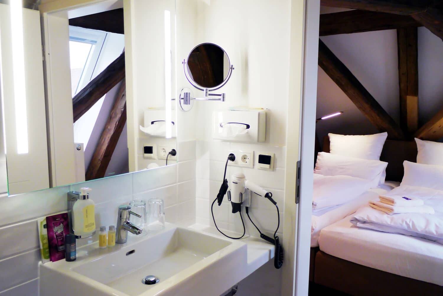 Ein kleines Badezimmer mit Waschbecken und Spiegelschrank, der Blick durch die offene Tür fällt auf ein Doppelbett.