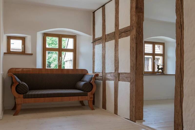 Impressionen aus einem der Dorf-Ferienhäuser: eine historische Couch vor einer rustikal erhaltenen Wand, die mit Holzbalken durchzogen ist.