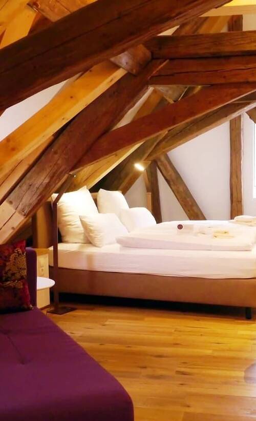 Gemütliches Schlafzimmer im Dachgeschoss mit sichtbaren Holzbalken, einem Doppelbett mit weißer Bettwäsche und einem violetten Sofa im Vordergrund.
