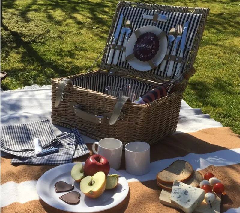 A wooden picnic basket on a brown and white striped picnic blanket covered with apples, bread, cheese, tomatoes, chocolate and crockery.