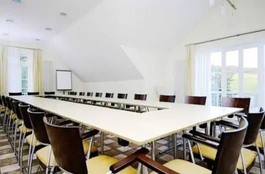 Ein großer Konferenztisch in U-Form mit weißem unbedeckten Tisch und schwarzen Tagungsstühlen in einem großen, hellen Raum.