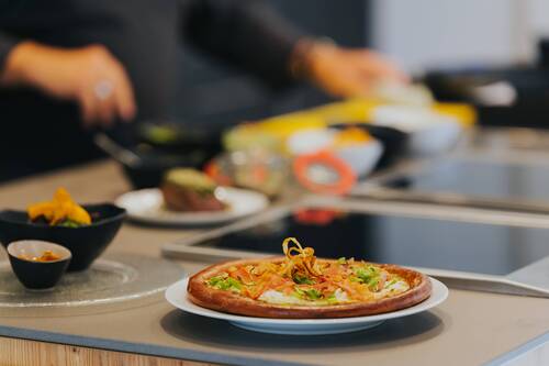 Frisch zubereitete Pizza auf einem Teller in moderner Showküche, während im Hintergrund Zutaten vorbereitet werden und gemeinsames Kochen zum Erlebnis wird.