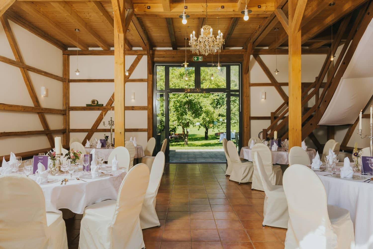 Elegant, rustic room with festive white tables and chandeliers, the view is directed towards the large patio door to the garden, through which you can see the green terrace - a wonderful conference and event location in Bavaria.