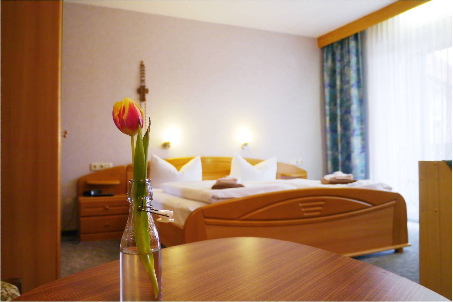 Ein Doppelzimmer im Landgathof Zum Hirschen mit Blick auf das Bett, im Vordergrund eine Flasche mit einer rot-gelben Tulpe im Fokus.