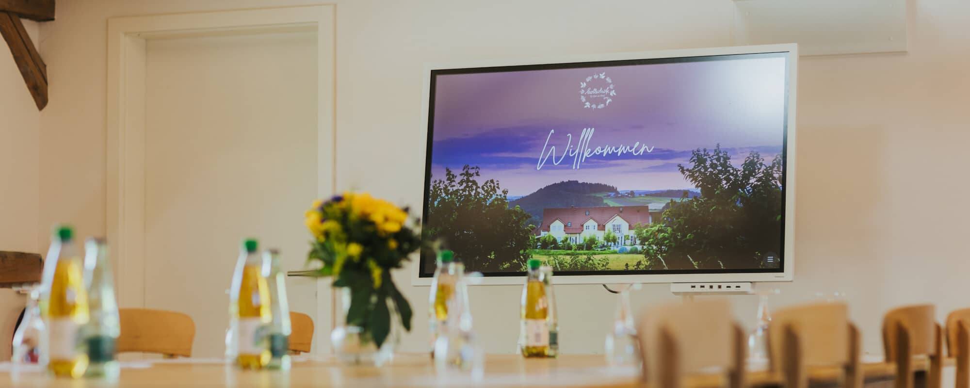 Conference room with wooden chairs, apple spritzer and water on the table and a modern presentation screen displaying a ‘Welcome’ sign as a location for conferences and seminars at the Hollerhöfe conference hotel.