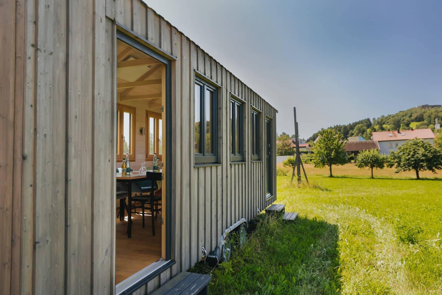 The entrance to the Tiny House photographed at an angle with a view of nature behind it. From the outside you can see the inside of the conference room on the left, looking into the distance you can see meadows, nature and the buildings of the Hollerhöfe far behind.