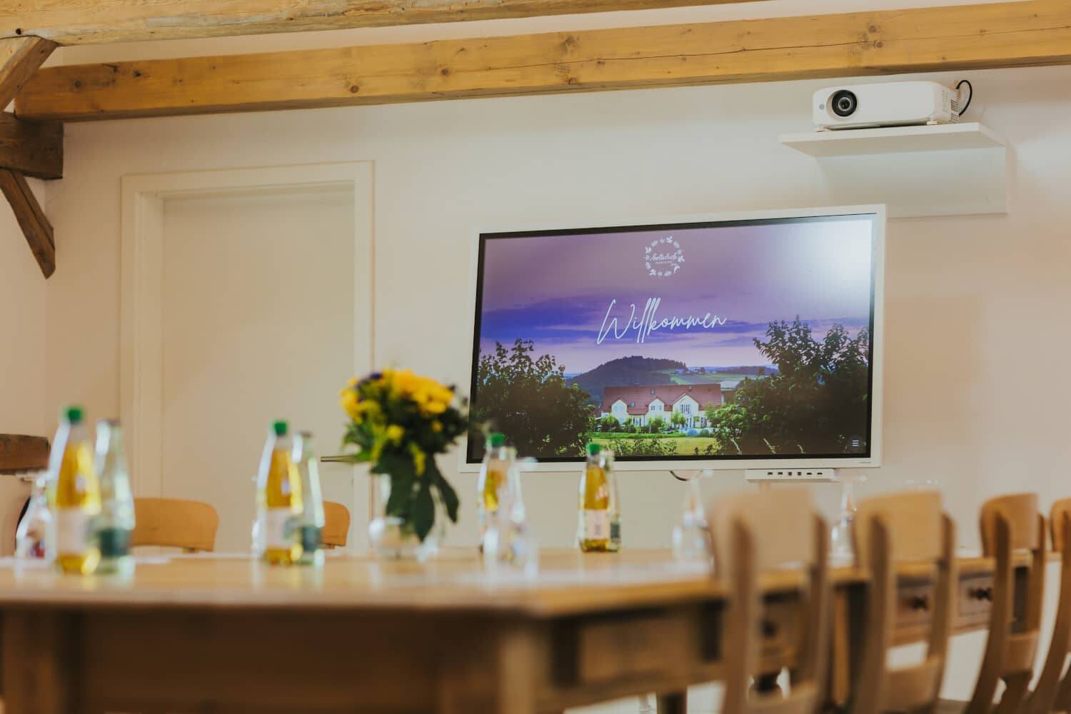 A picture focussing on the conference TV in the Hollermanufaktur with the Hollerhöfe welcome screen. The table with the conference drinks can be seen out of focus in the foreground.