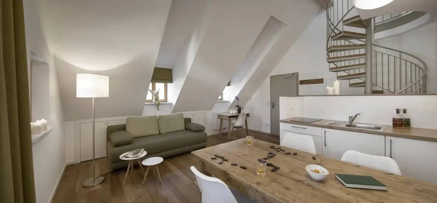 Der Wohnraum im Deluxe Apartment mit rustikalem Esstisch, auf dem gerade ein Domino Spiel liegt, einer Küchenzeile dahinter und einer grünen Zweisitzer-Couch.