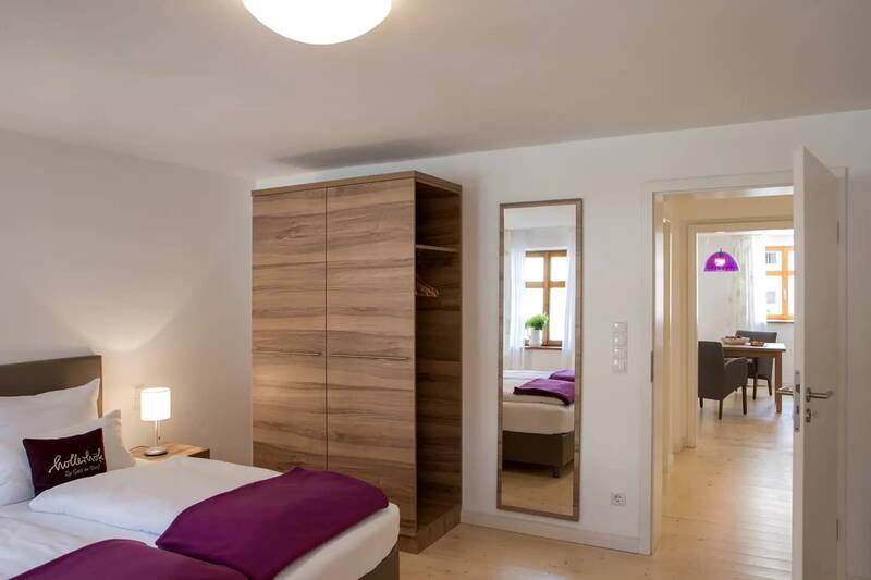 Ein Schlafzimmer mit Bett, Schrank und Spiegel. Der Blick fällt durch die geöffnete Zimmertür in den Wohn- und Essbereich.
