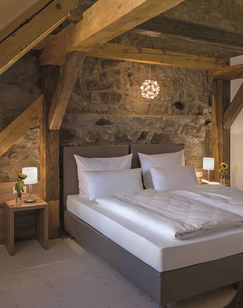 Cozy bedroom with rustic charm, featuring a double bed with white bedding and a gray bed frame, flanked by two nightstands with small lamps and flower vases. The walls are made of exposed natural stone, and the ceiling is supported by heavy wooden beams. A decorative hanging lamp provides warm lighting.