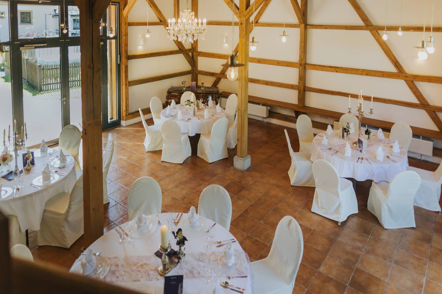 Elegant, rustic ballroom with white-clothed tables, chandeliers and a large window front, easily photographed from an elevation - the Hollerscheune as a conference and event location.
