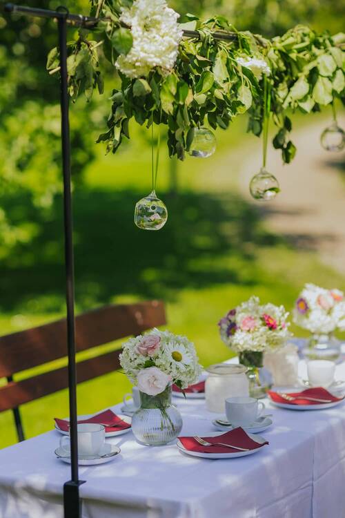 Liebevoll gedeckter Tisch im Grünen mit Blumenarrangements und hängender Dekoration – die Hollerhöfe als perfekte Location für eine Gartenparty.