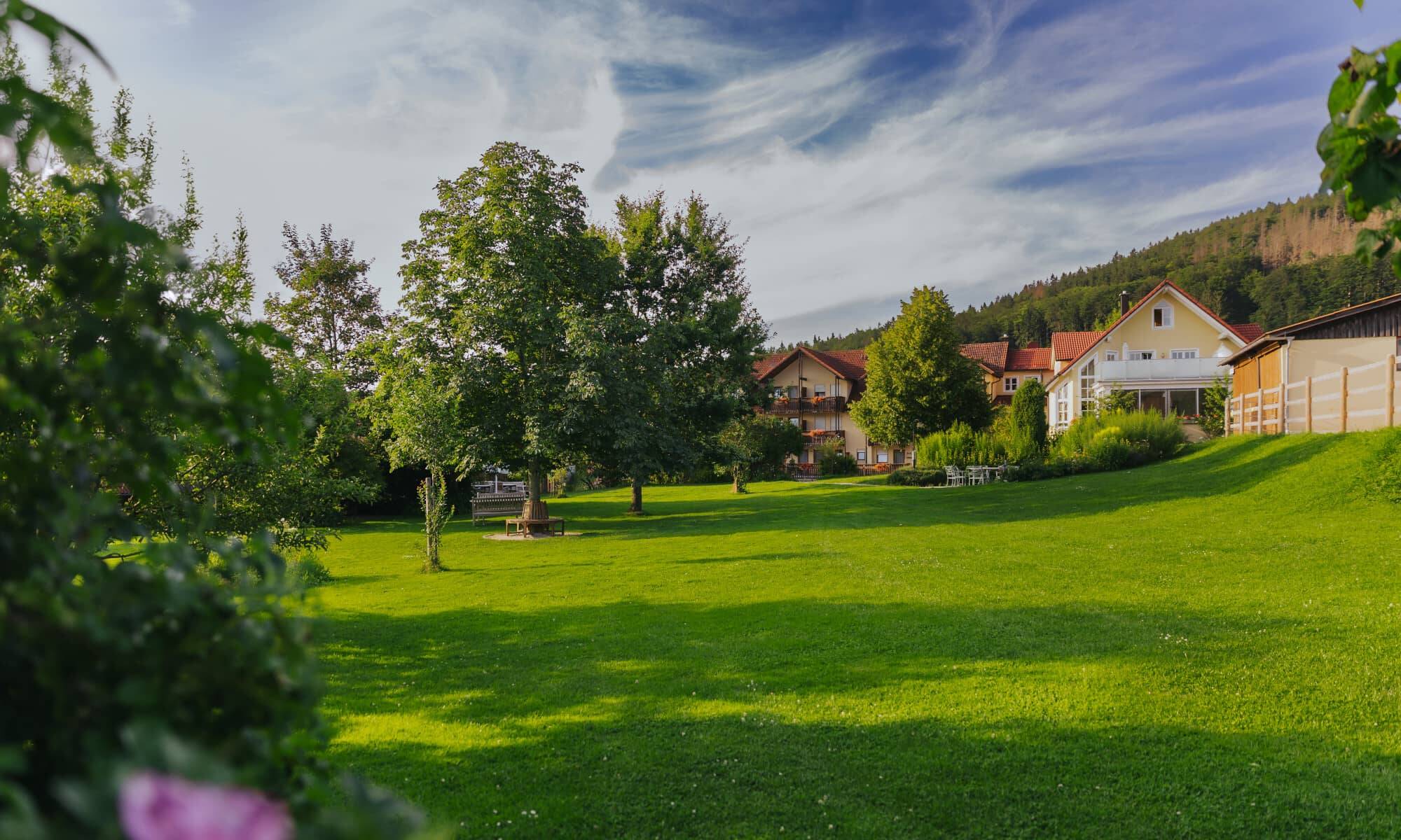 Der Garten vom Hotel Hollerhöfe mit weiter grüner Wiese, sanften Hügeln und Bäumen. Im Hintergrund sieht man das Haupthaus und darüber den blauen Himmel mit weißen Kondensstreifen.