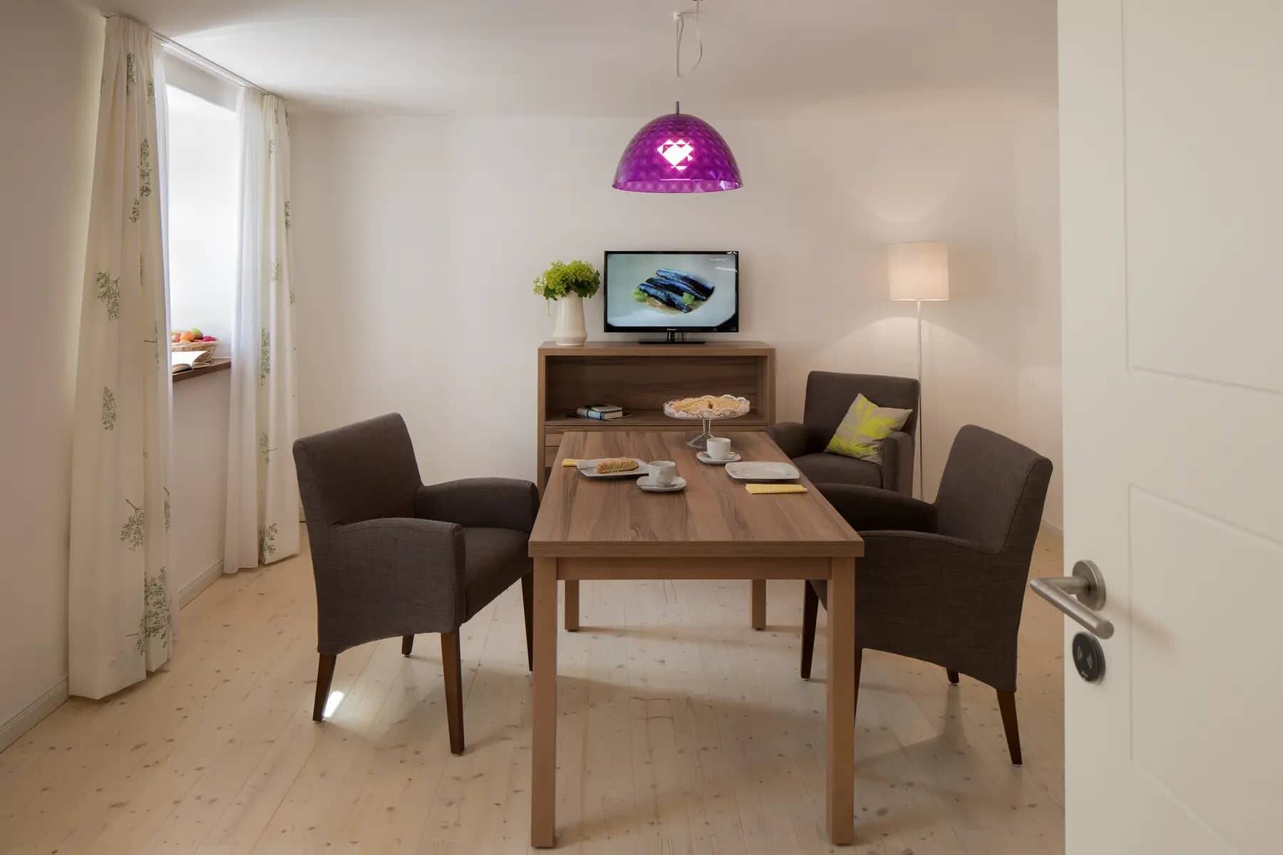 Ein Holztisch im Esszimmer der Ferienwohnung Hollerhöfe mit drei gemütlichen Stühlen, gedecktem Tafelservice und einem Fernseher auf einem Regal dahinter.