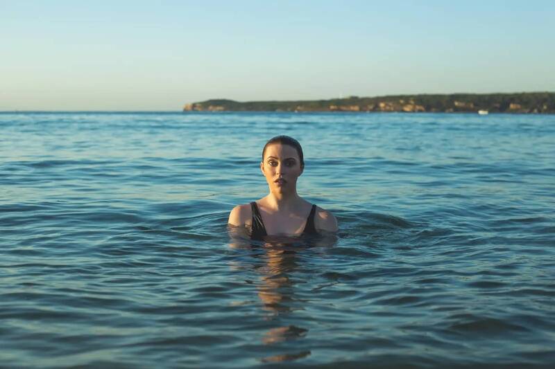 A dark-haired woman in a black swimming costume in blue water, only her upper body slightly protruding from the water.