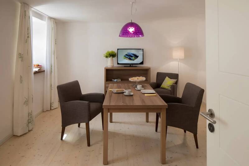 Der Esstisch in einem der Dorfhäuser der Hollerhöfe: ein moderner Holztisch mit 3 Stühlen, einem Regal mit Fernseher oben drauf.