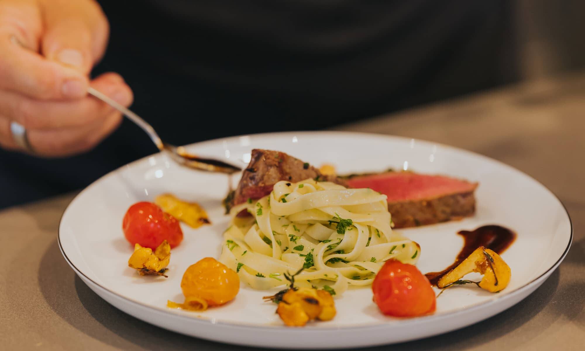 Person genießt ein feines Gericht mit zartem Fleisch, Tagliatelle und Cherrytomaten – ein Beispiel für die kulinarischen Erlebnisse, die bei einem Kochkurs in den Hollerhöfen zubereitet werden können.